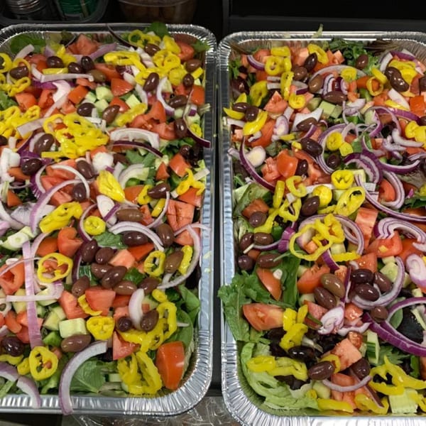 Two large catering trays filled with fresh garden salad, topped with olives, tomatoes, and peppers.