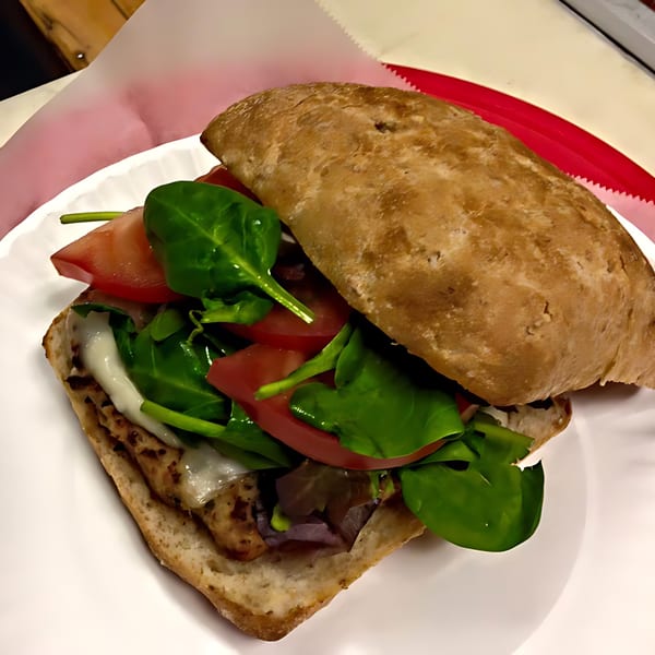 A chicken sandwich on ciabatta bread with melted cheese, spinach, and tomatoes on a white plate.