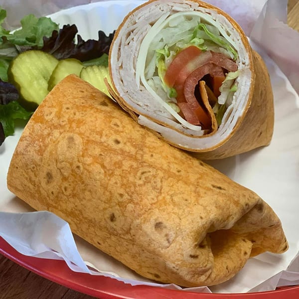 A turkey wrap, cut in half, revealing lettuce, tomato, and cheese, served with pickles and salad.