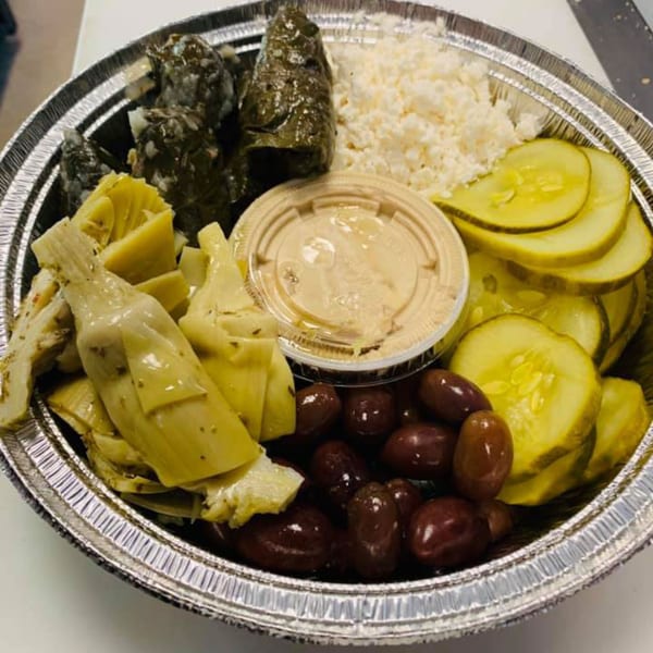 A foil takeout container filled with dolmades, feta, artichoke hearts, pickles, olives, and hummus.
