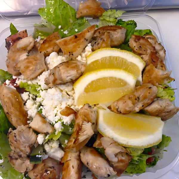 Chicken salad with crumbled feta cheese, lemon slices, and fresh greens in a clear container.