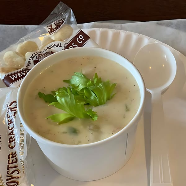 A white bowl of creamy clam chowder topped with fresh parsley, alongside oyster crackers and a spoon.