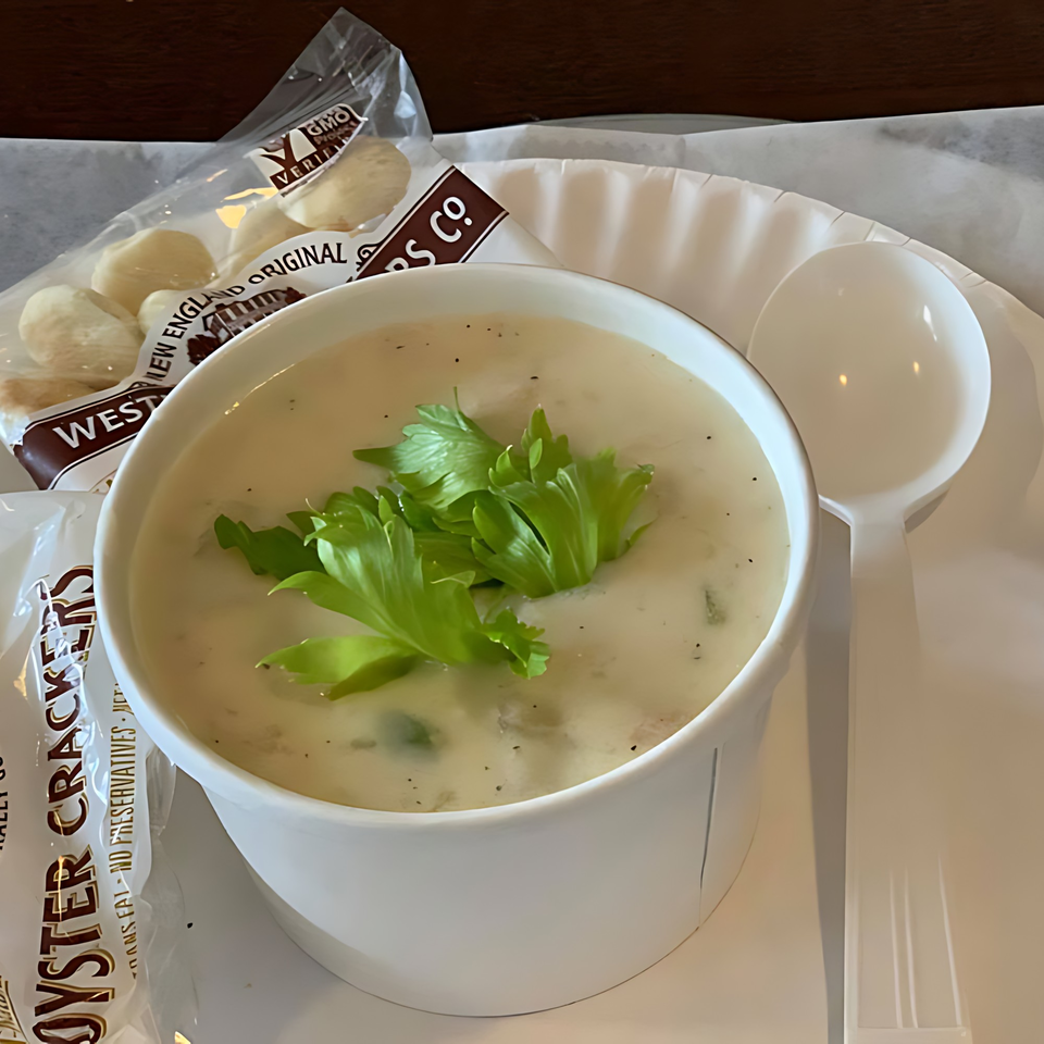 A white bowl of creamy clam chowder topped with fresh parsley, alongside oyster crackers and a spoon.