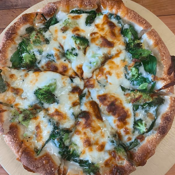 A freshly baked pizza from The Original Village Pizza, topped with melted cheese, broccoli, and spinach.