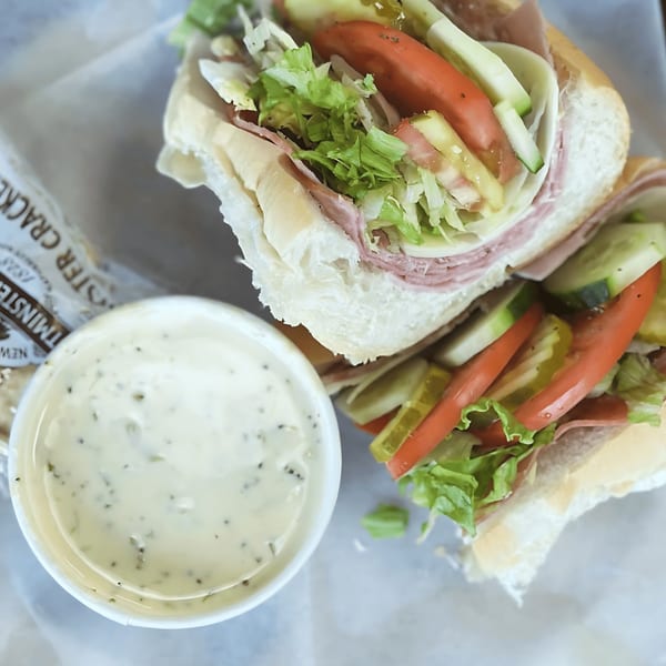 A large deli sub sandwich with meat, cheese, lettuce, tomatoes, and pickles, next to a cup of dressing.